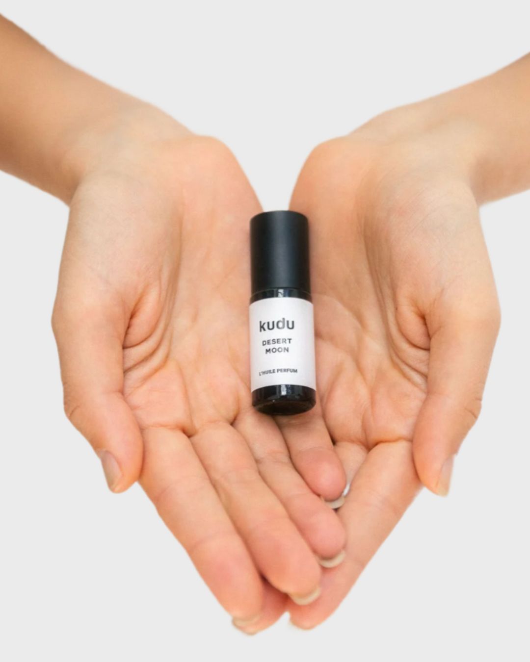 Roll-on perfume titled "Kudu Desert Moon." Two hands holding the amber bottle with a black cap, showcasing a sleek, minimalist design on a white background.