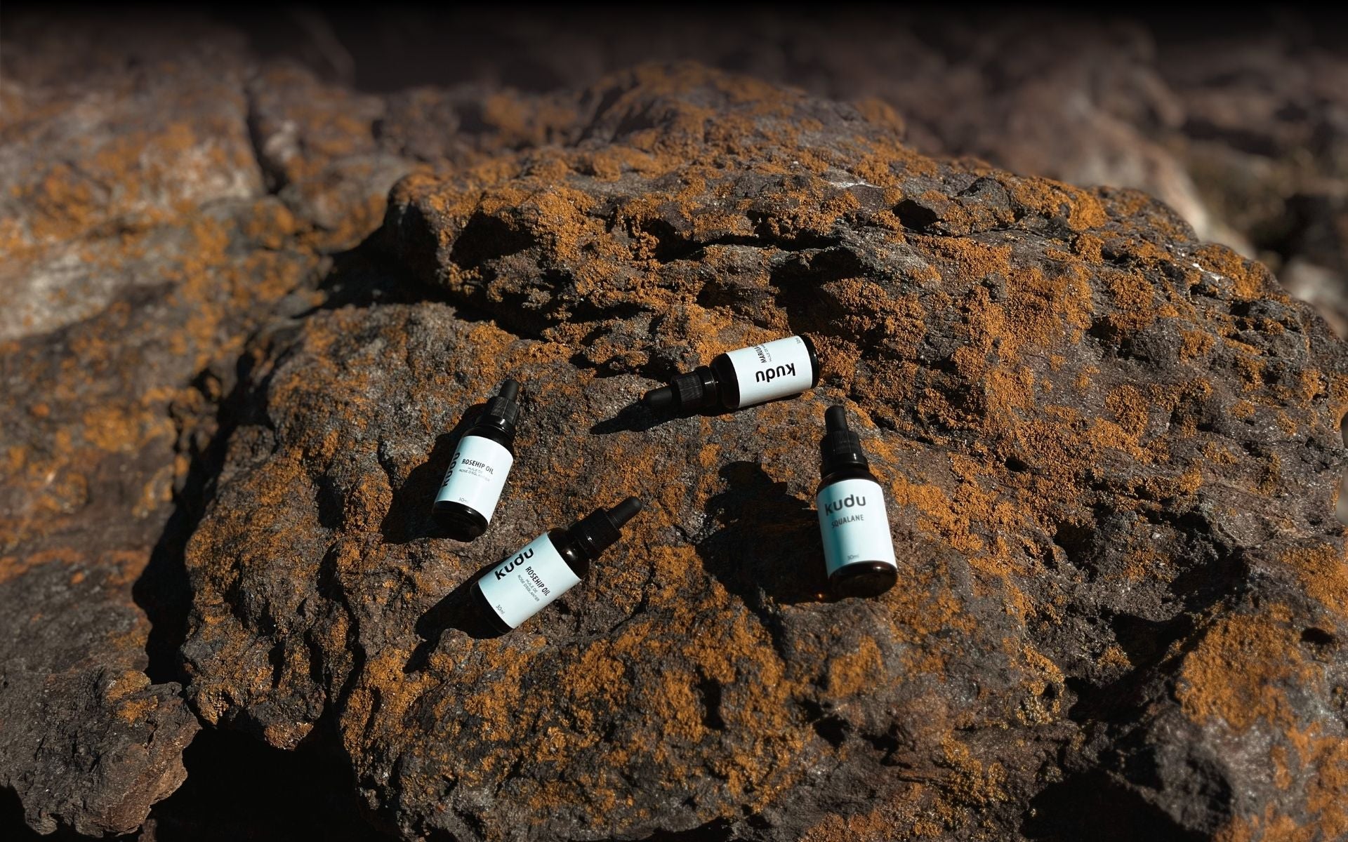 Bottles of skincare oil labeled "kudu" are scattered on a sunlit, lichen-covered rock. The scene conveys a sense of natural beauty and tranquility.