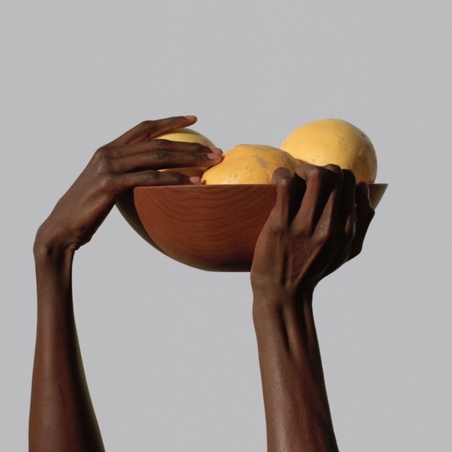 Hands holding a wooden bowl containing three large marula fruits against a plain gray background.